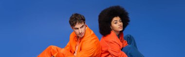 African american model looking at camera near man in orange clothes isolated on blue, banner 