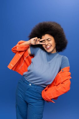 Cheerful african american woman in bright outfit showing peace sign on blue background