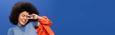 Cheerful african american woman in stylish outfit gesturing on blue background, banner 