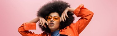 Stylish african american model in orange jacket and sunglasses isolated on pink, banner 