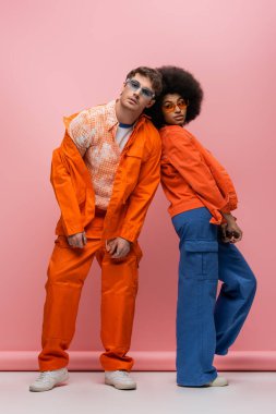 Stylish interracial couple in jackets looking at camera on pink background 