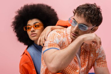 Portrait of brunette man in sunglasses posing near african american woman isolated on pink 