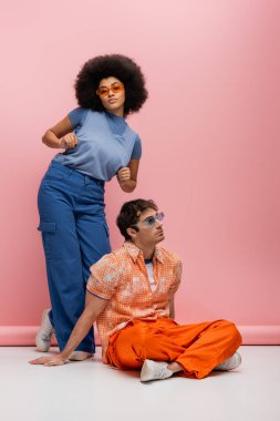 Fashionable african american woman in sunglasses and blue clothes posing near man on pink background 