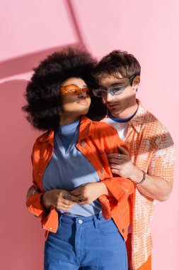 Stylish man in sunglasses hugging african american woman on pink background with shadow 