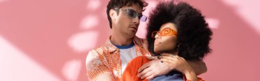 Stylish man in sunglasses hugging and looking at african american woman on pink background with shadow, banner 