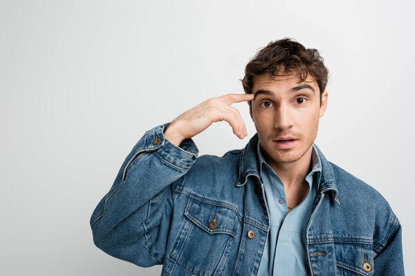 overwhelmed man in denim jacket pointing with finger at head and looking at camera isolated on grey 