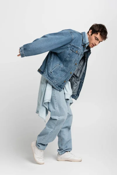 full length of stylish man in denim jacket and jeans posing while looking down on grey