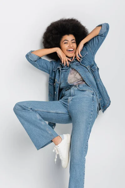 Cheerful african american woman in jeans and denim vest standing on grey background
