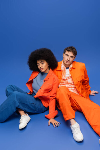 Multiethnic couple in orange jackets sitting on blue background