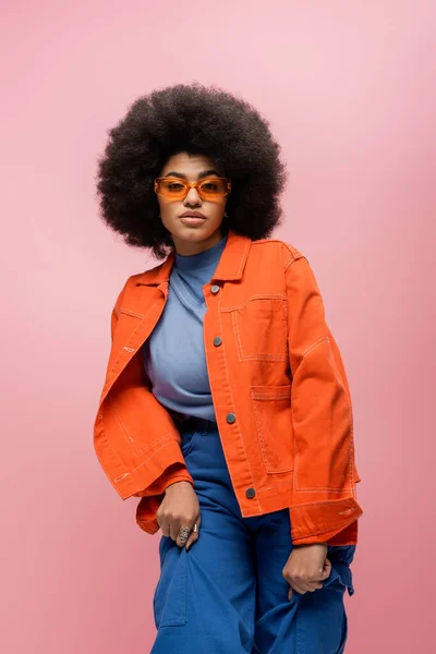 Curly african american model in orange jacket and sunglasses isolated on pink