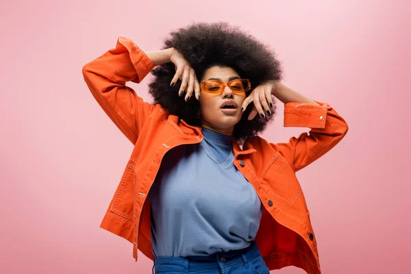 Trendy african american model in orange jacket and sunglasses posing isolated on pink