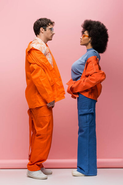 Вид сбоку трезвенной межрасовой пары в солнцезащитных очках, смотрящей друг на друга на розовом фоне 