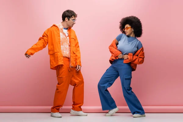 stock image Full length of stylish multiethnic couple in sunglasses and bright jackets posing on pink background 