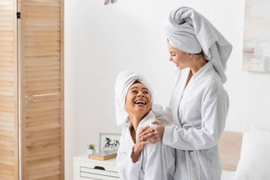 laughing african american woman in white towel and bathrobe looking at smiling friend in bedroom