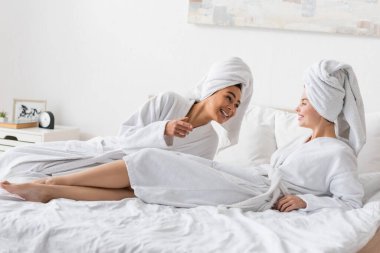 cheerful african american woman talking to pretty friend lying on bed in white terry bathrobe and towel 