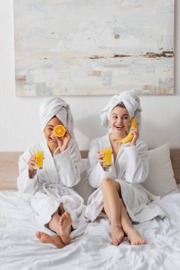 full length of cheerful interracial women with fruits and orange juice having fun while sitting on bed