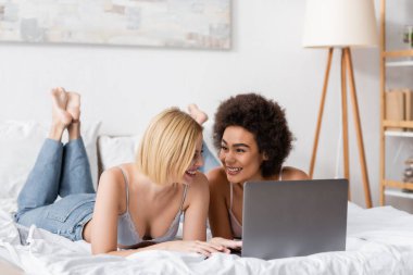 blonde woman in bra and jeans using laptop near cheerful african american friend on bed