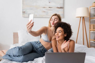 blonde woman pouting lips while taking selfie with smiling african american friend waving hand in bedroom