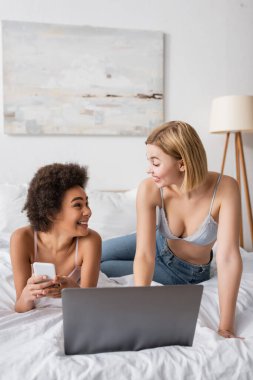 young interracial women in bras and jeans smiling at each other near laptop in bedroom