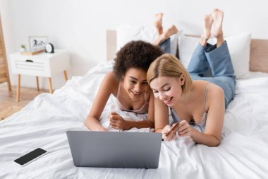 young blonde woman holding credit card near african american friend and laptop in bedroom clipart