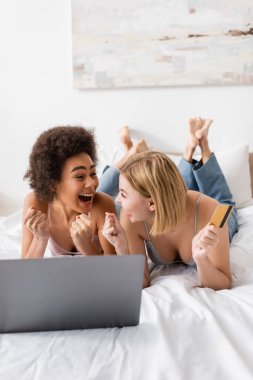 excited multiethnic friends looking at each other and showing win gesture near credit card and laptop on bed at home