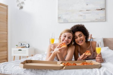 joyful multiethnic women looking at camera while lying on bed with delicious pizza and champagne glasses with cocktails