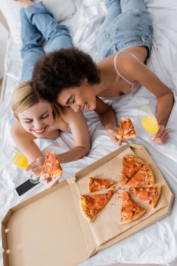 high angle view of interracial women smiling with closed eyes near tasty pizza and cocktails in champagne glasses while lying on bed clipart