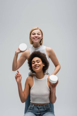 young interracial women in white tank tops posing with body cream and smiling at camera isolated on grey clipart