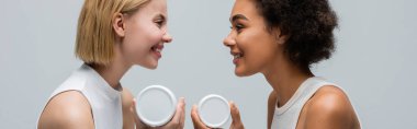 side view of interracial blonde and brunette women with body cream smiling at each other isolated on grey, banner