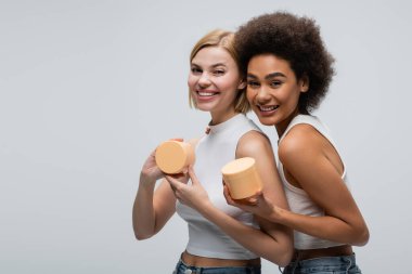 cheerful interracial models in white tank tops smiling at camera while holding cosmetic cream isolated on grey clipart