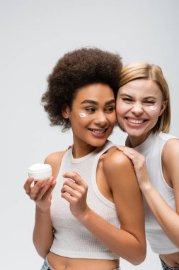 Afro-Amerikan modelinin yanındaki kameraya bakan neşeli sarışın kadın gri renkli yüz kremiyle poz veriyor.