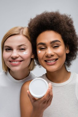 happy african american woman holding face cream near young blonde model isolated on grey clipart