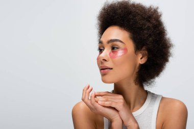 curly african american woman in hydrogel eye patches looking away isolated on grey
