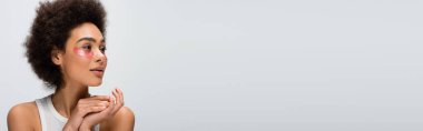 young and pretty african american woman in collagen eye patches looking away isolated on grey, banner
