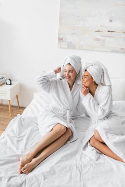 positive african american woman in white robe and towel talking to smiling friend looking away on bed
