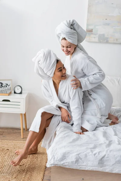 happy woman in white robe and towel embracing african american friend in bedroom