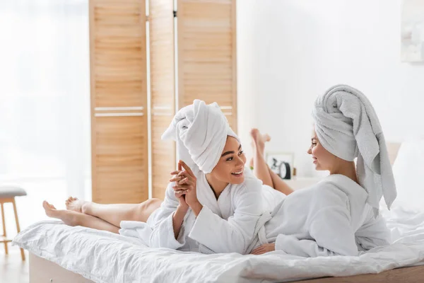 Щасливі Міжрасові Жінки Білих Яких Халатах Рушниках Розмовляють Відпочиваючи Ліжку — стокове фото