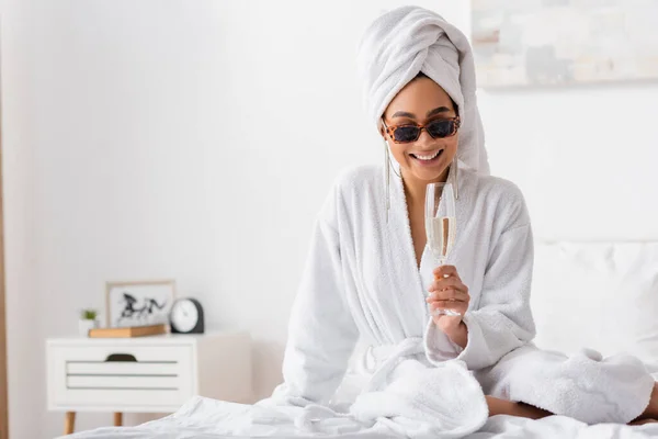 stock image happy african american woman in terry bathrobe and trendy sunglasses sitting on bed with glass of champagne