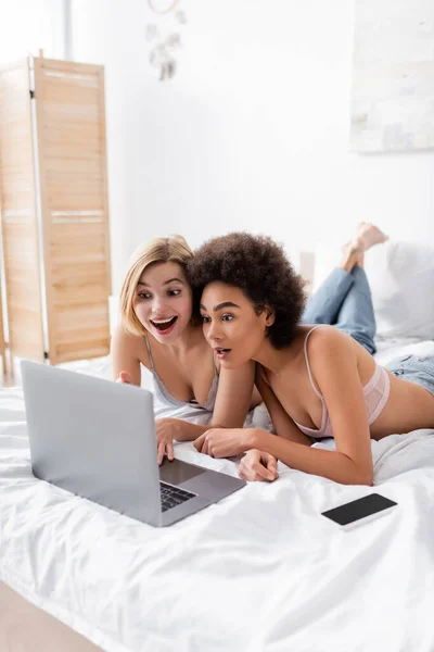 amazed blonde woman pointing at laptop near african american friend and smartphone with blank screen on bed