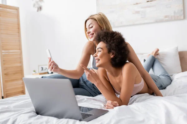 smiling african american woman pointing with finger near blonde friend with mobile phone and laptop on bed