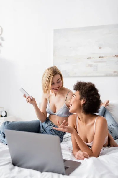 blonde woman holding smartphone near smiling african american friend pointing at laptop on bed