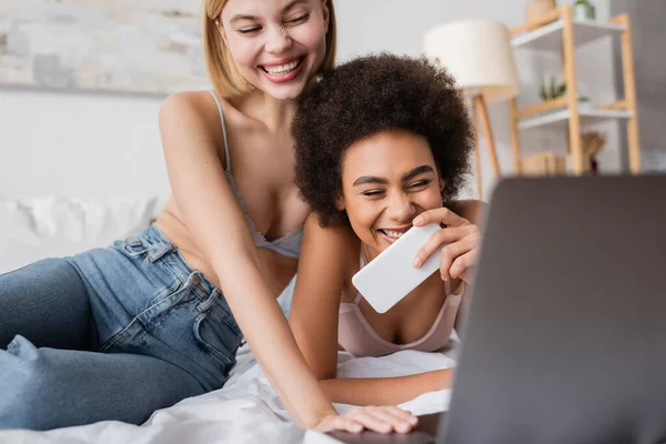 excited african american woman holding mobile phone and looking at blurred laptop near blonde friend