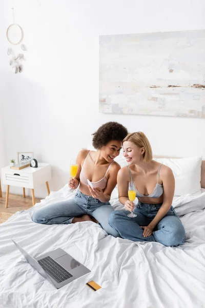 stock image happy african american woman showing smartphone to smiling friend while sitting with cocktail near laptop and credit card on bed