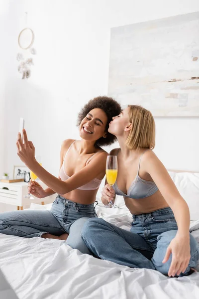blonde woman with cocktail in champagne glass kissing smiling african american friend taking selfie on bed at home