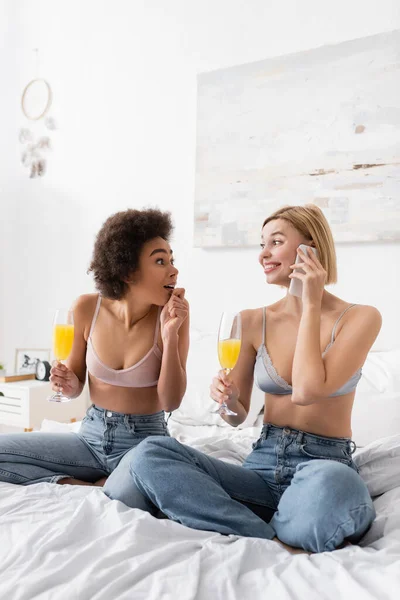 amazed african american woman with cocktail looking at blonde friend talking on mobile phone