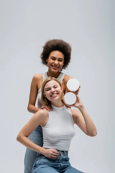 excited multiethnic models in white tops and jeans posing with body cream and looking at camera isolated on grey