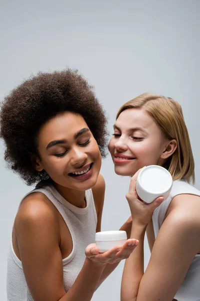 happy african american woman looking at container with body cream near blonde model isolated on grey