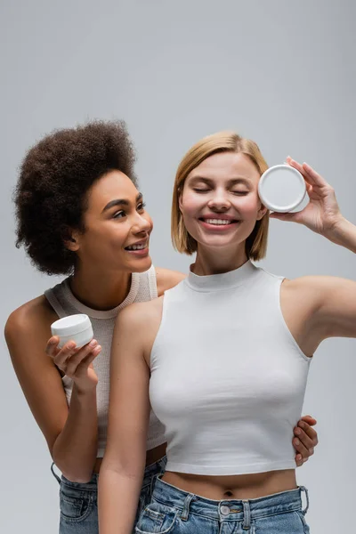 happy blonde model with closed eyes posing with body cream near african american woman isolated on grey