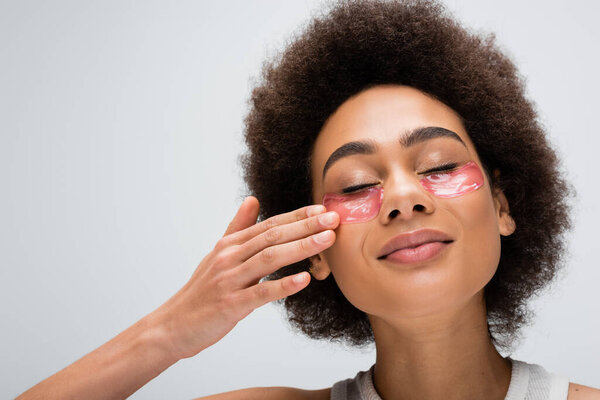 pleased african american woman with closed eyes touching hydrogel eye patch isolated on grey