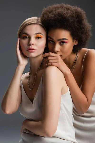 stock image pretty multiethnic women with colorful visage posing in white strap dresses and looking at camera isolated on grey